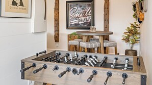 Una habitación con mesa de fútbolín, un estante de madera montado en la pared, una cita inspiradora enmarcada, una pequeña mesa redonda con sillas y una planta en maceta.