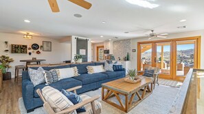 A spacious living room with a large blue sofa, wooden coffee table, and a view of a hilly landscape through large sliding doors. - Hilltop Hacienda by Avantstay Game Room + Hot Tub Hilltop Paso Views (Paso Robles)