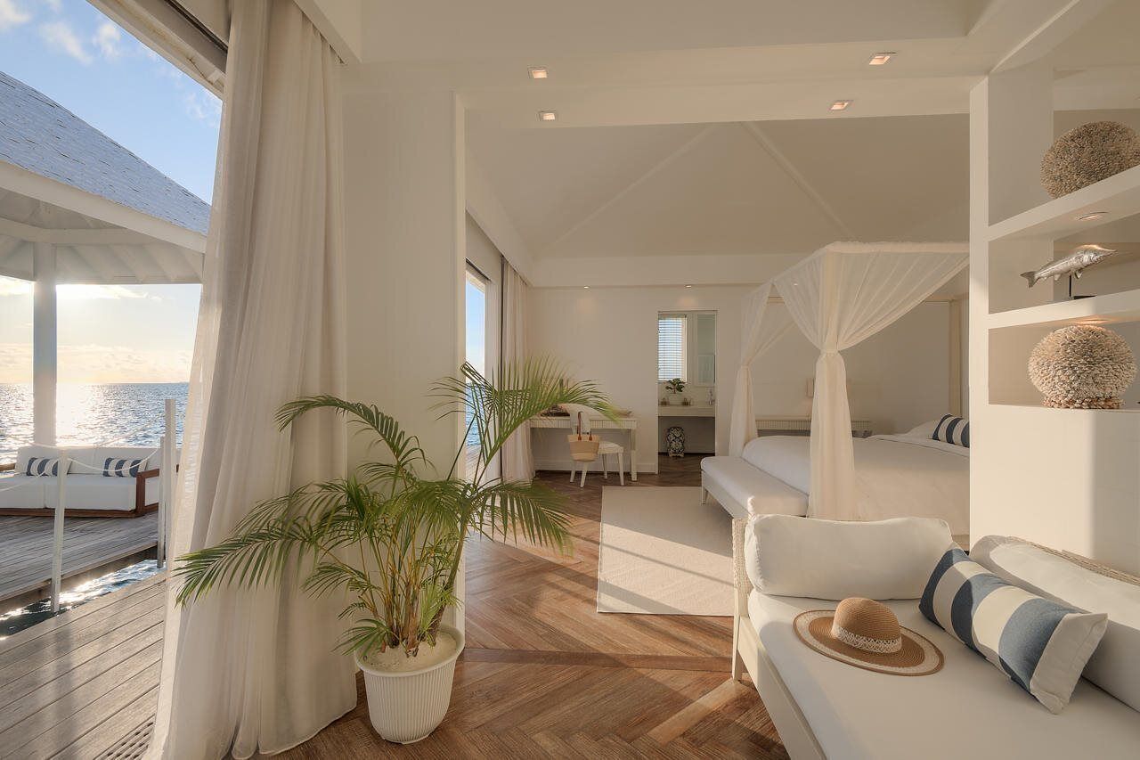 Habitación de hotel moderna con piso de madera, cama con dosel, un espacio de descanso con sofá y sombrero, comedor con mesa y sillas, y un amplio ventanal con vista al mar.