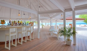 A covered outdoor bar area with a wooden floor, white bar stools, and a variety of bottles. - Diamonds Athuruga Maldives Resort and SPA (Athuruga Island)