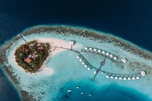 An aerial view of a resort island with a central building surrounded by trees, a beach, and a crescent-shaped lounge area with umbrellas.