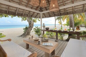 A wooden deck with a table set for dining, overlooking a beach with palm trees and the ocean.