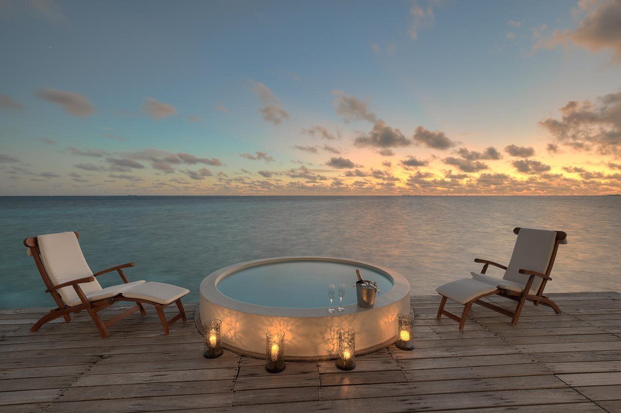 Una terraza de madera con un jacuzzi circular rodeado por dos sillones de descanso, con vista al mar tranquilo al atardecer.