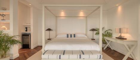 A four-poster bed with a white canopy, a bedside table with a lamp, a small potted plant, and a white console table with a lamp.
