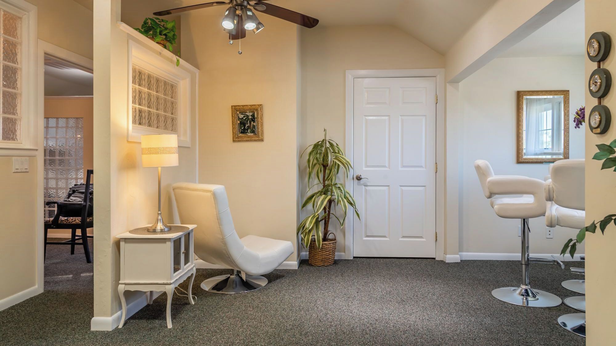 A hallway with a white door, a ceiling fan, a lamp, a chair, a potted plant, and a mirror on the wall.