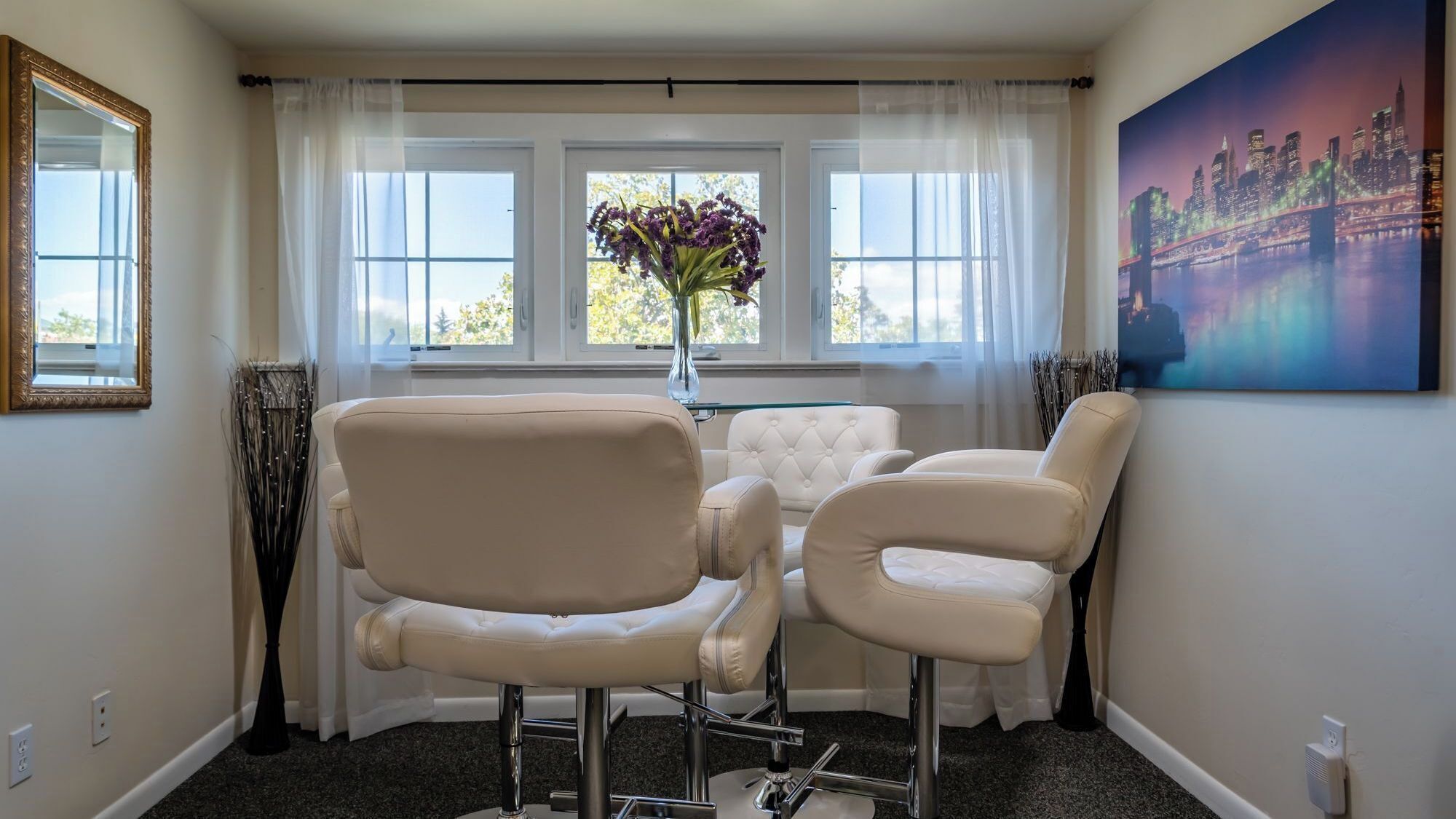 A room with three white leather chairs, a vase with purple flowers, a painting of a cityscape, and a window with white curtains.