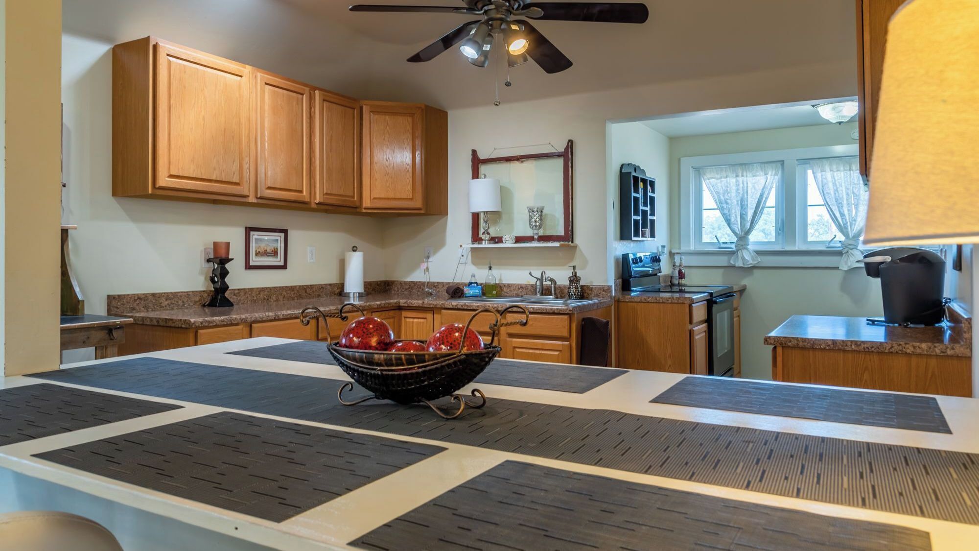 A kitchen with wooden cabinets, a central island with a bowl of fruit, a ceiling fan, and a window with curtains.