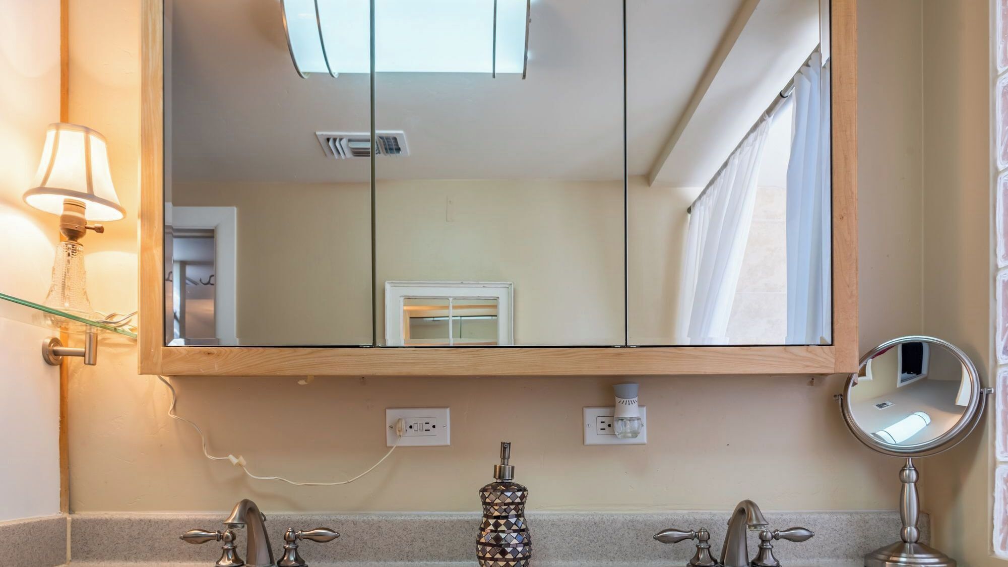 A bathroom with a large mirror, two faucets, a glass soap dispenser, and a wall-mounted light fixture.