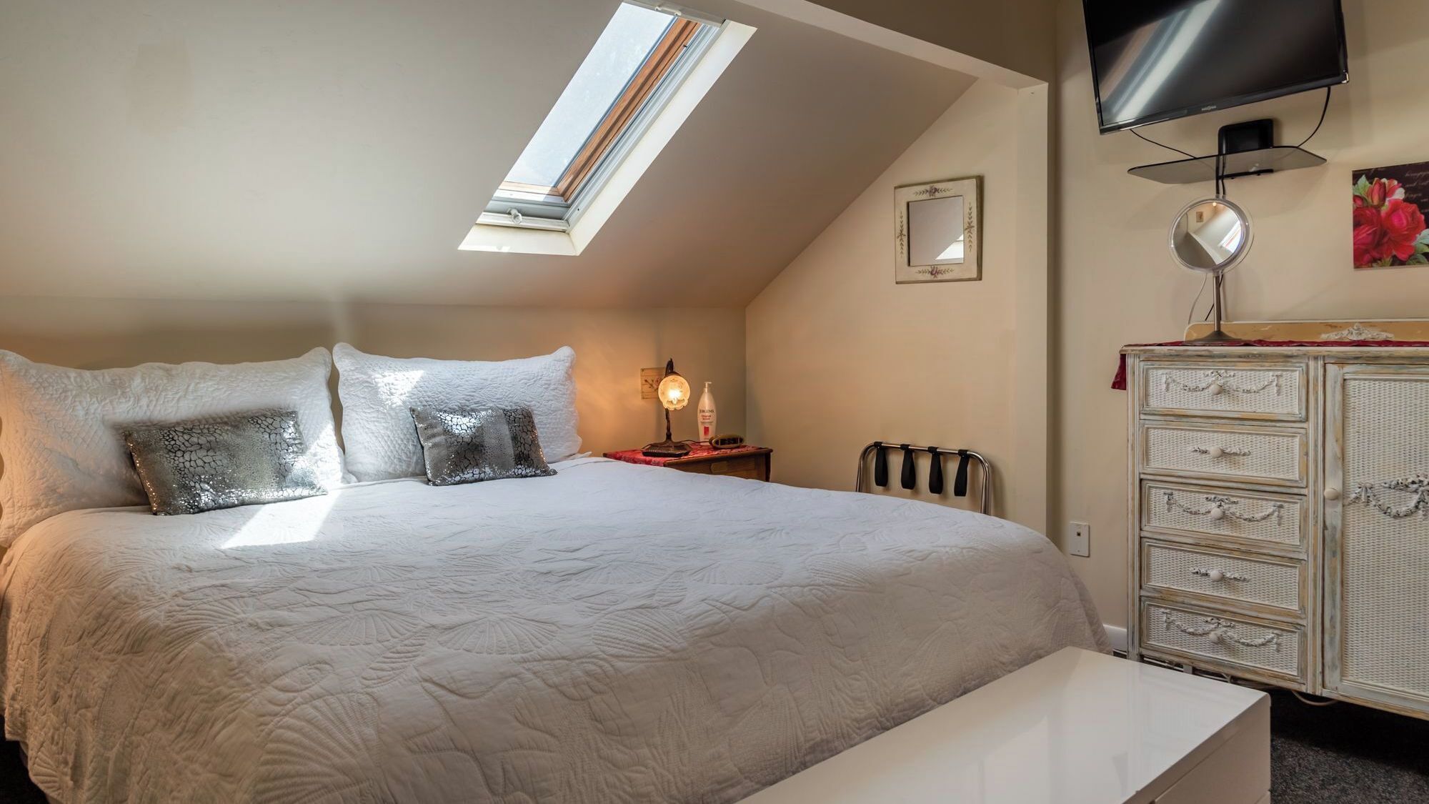 A bedroom with a bed, a bedside table, a lamp, a dresser, a television, and a skylight.