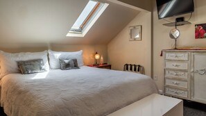A bedroom with a bed, a bedside table, a lamp, a dresser, a television, and a skylight.
