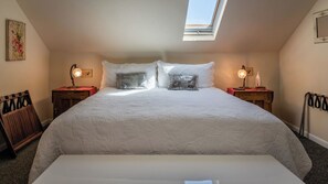 A bedroom with a large bed, two bedside tables with lamps, a chair, a skylight, and a wall-mounted picture.