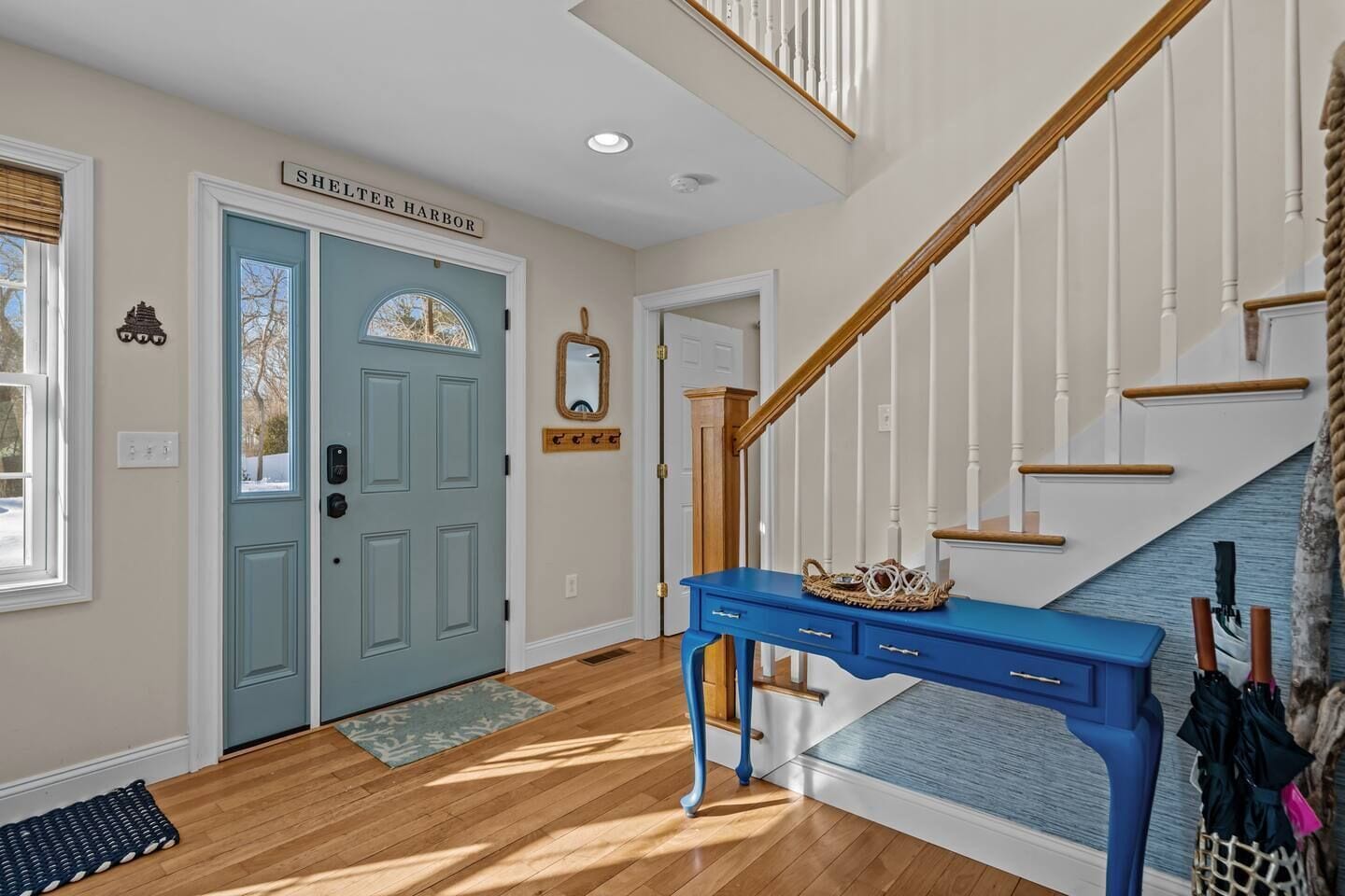 A hallway with a blue door labeled 'Shelter Harbor,' a wooden staircase, and a blue console table with a basket.