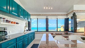 Uma cozinha com vista para o oceano, com armários cor turquesa, bancadas de granito e uma área de jantar também com vista.