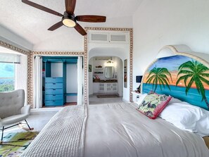 A bedroom with a bed, a ceiling fan, a view of the bathroom, and a painting on the wall. - Penthouse Paradise | Wraparound Ocean Views (Isla Mujeres)