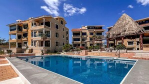 Piscina cercada por edifícios altos com varandas, uma construção com telhado de palha e um céu azul claro.