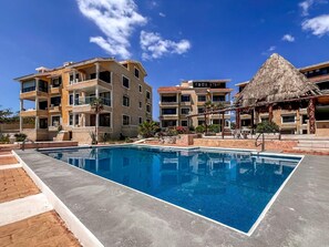 A swimming pool surrounded by multi-story buildings with balconies, a thatched-roof structure, and a clear blue sky. - Penthouse Paradise | Wraparound Ocean Views (Isla Mujeres)
