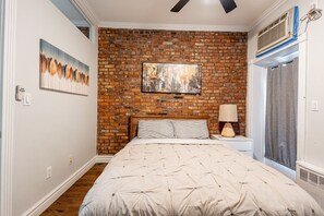 Un dormitorio con pared de ladrillo, una cama con almohada a rayas, un cuadro y un ventilador de techo.