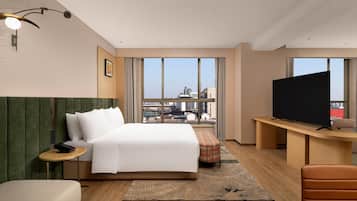 Habitación de hotel con cama, escritorio con televisor, silla, mesita y vistas a la ciudad a través de amplias ventanas.