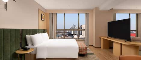 Habitación de hotel con cama, escritorio con televisor, silla, mesita y vistas a la ciudad a través de amplias ventanas.