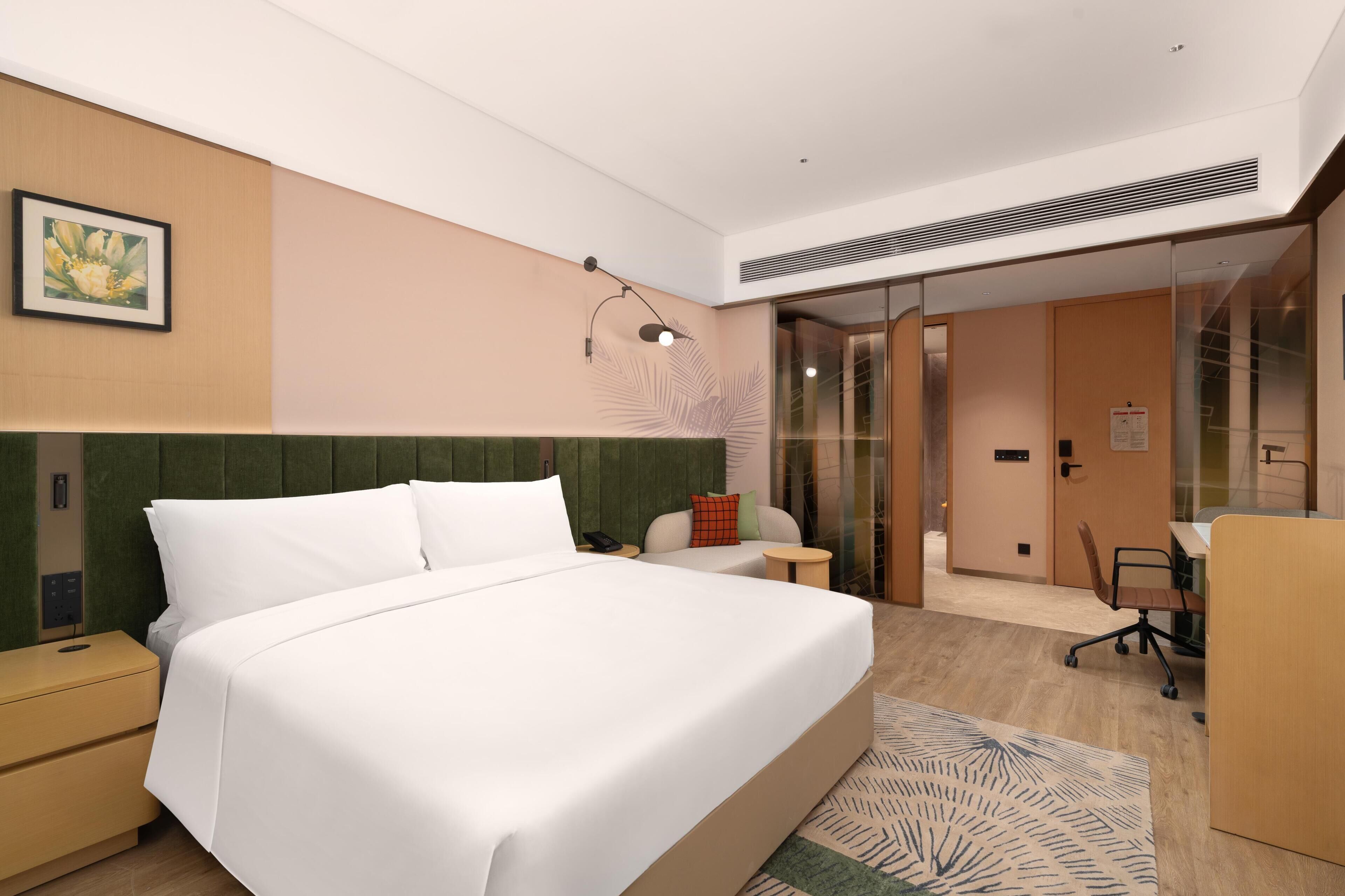 A hotel room with a bed, bedside table, a desk with a chair, a glass door leading to a bathroom, and a wall-mounted artwork.