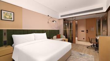 A hotel room with a bed, bedside table, a desk with a chair, a glass door leading to a bathroom, and a wall-mounted artwork.