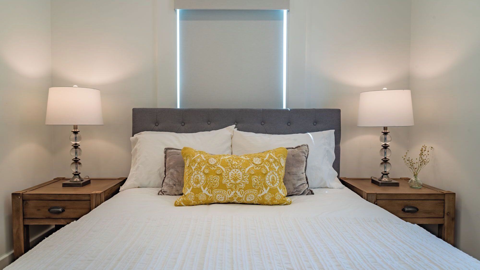 Una cama bien hecha con un cabecero gris, dos mesitas de noche con lámparas y una ventana sobre la cama.