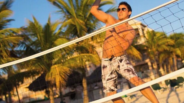 Eine Person spielt Beachvolleyball auf einem Sandplatz, im Hintergrund sind Palmen zu sehen.