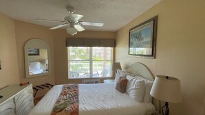 Ein Hotelzimmer mit Bett, Deckenventilator, Fenster mit Jalousien, Nachttisch mit Lampe und Kommode.