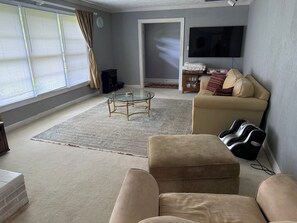 A living room with a beige sofa, a glass coffee table, a television mounted on the wall, and a large window with blinds. - Tranquil 3-bedroom house with AC in charming Spruce Pine (Spruce Pine)