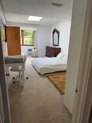 Ein Schlafzimmer mit Bett, Schminktisch, Schreibtisch mit Stuhl, Heizkörper und einem Fenster mit Blick ins Grüne.
