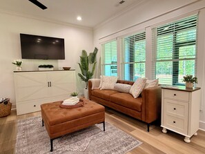 A living room with a brown sofa, a white cabinet, a flat-screen TV, and a large window with blinds. - Refuge at Augusta 12 Min to LU  Quiet Community with private Screened Porch (Rustburg)