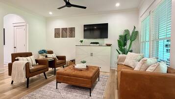 A modern living room with a brown sofa, a brown armchair, a coffee table, a ceiling fan, a flat-screen TV, and a large window with blinds.