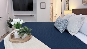 A bedroom with a bed, a TV mounted on the wall, a wooden tray with flowers, and a door leading to a bathroom. - Refuge at Augusta 12 Min to LU  Quiet Community with private Screened Porch (Rustburg)