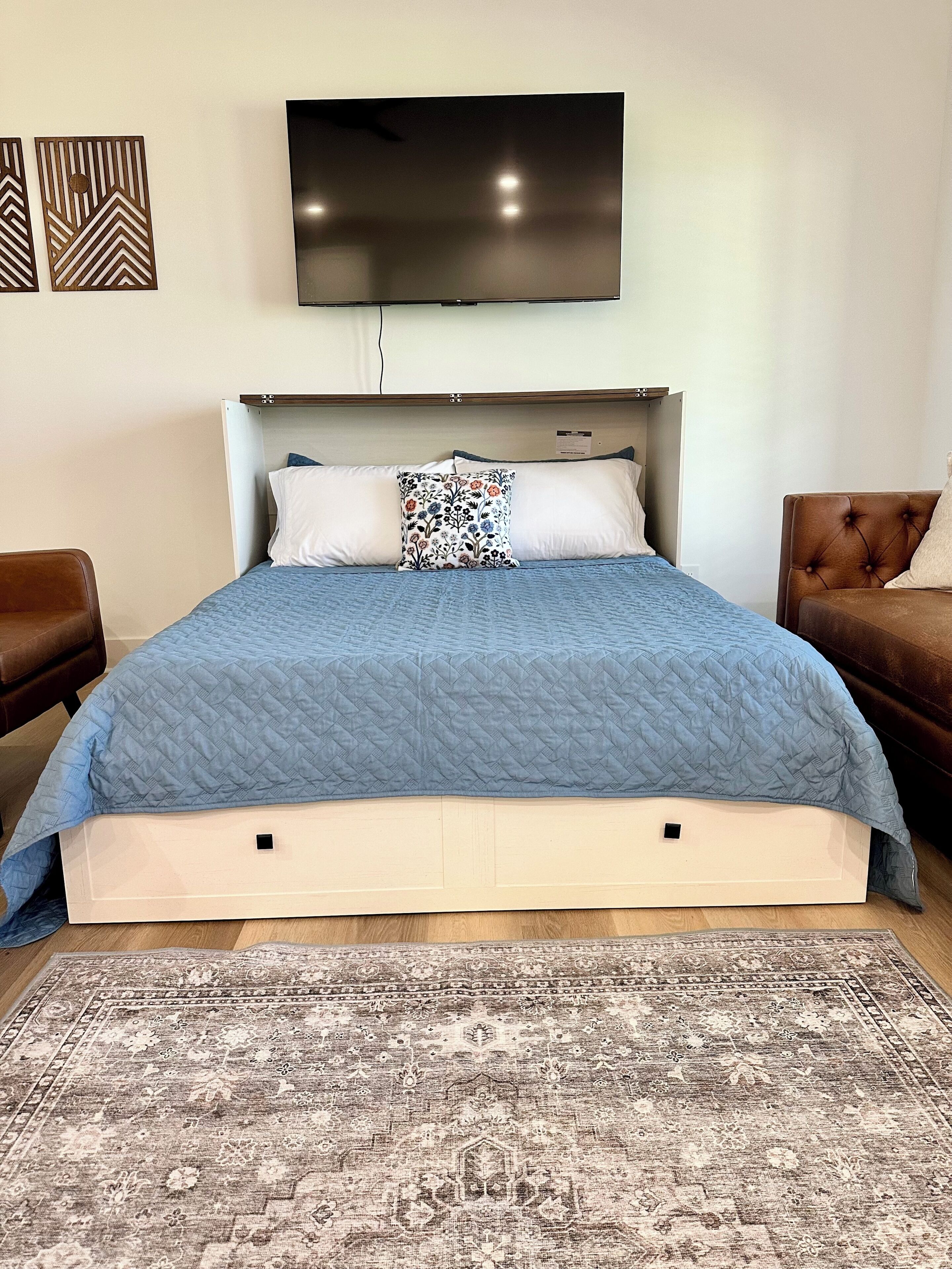 A neatly made bed with a blue quilt, a patterned pillow, and a flat-screen TV mounted above it.