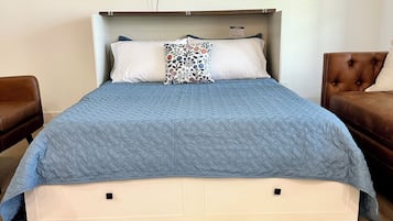 A neatly made bed with a blue quilt, a patterned pillow, and a flat-screen TV mounted above it.