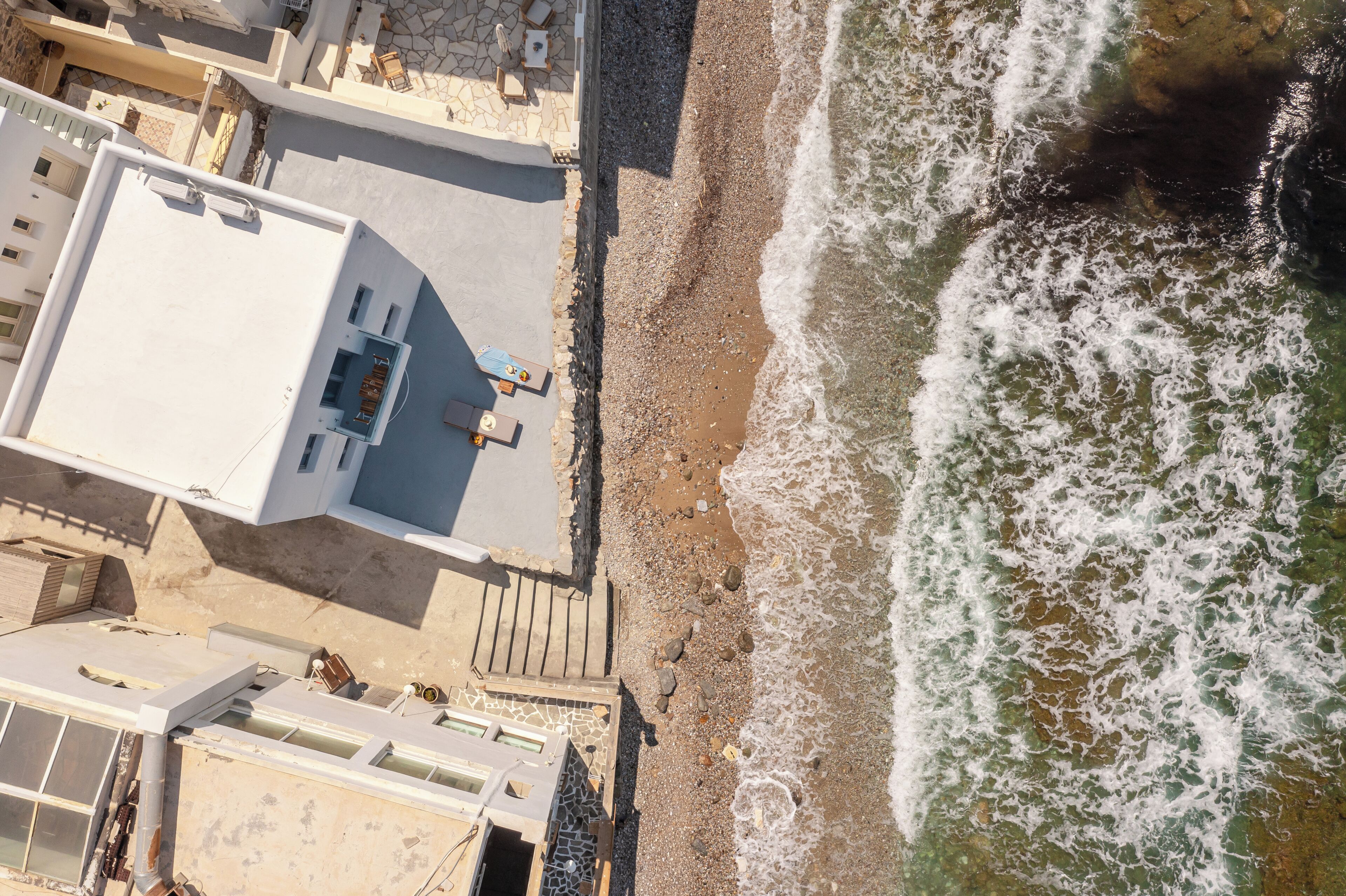 Vista aérea de un edificio costero con terraza en la azotea, junto a una playa de arena y olas.
