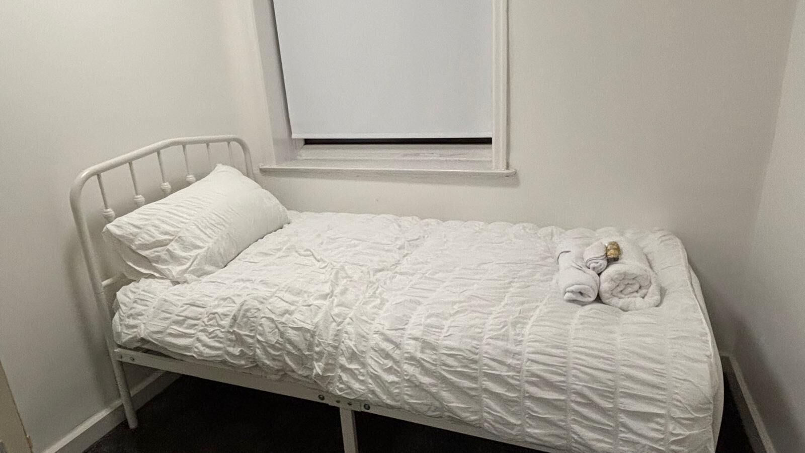 A single-bed room with a white metal frame, a window with closed blinds, and a white bedspread.