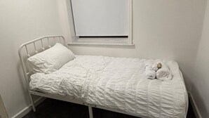 Habitación con cama individual de estructura metálica blanca, una ventana con persianas cerradas y una colcha blanca.