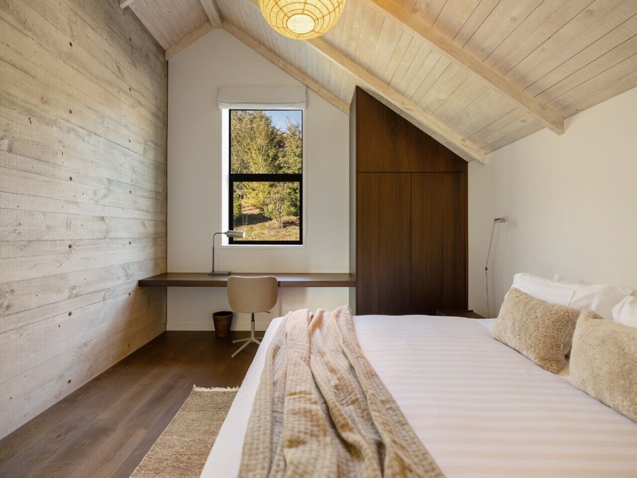 Een slaapkamer met een schuine plafond, een groot raam, een bed met kussens, een bureau en een stoel.