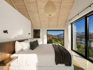 Ein modernes Schlafzimmer mit einem großen Bett, einem Holz-Kopfteil und Blick auf eine gebirgige Landschaft durch raumhohe Fenster.