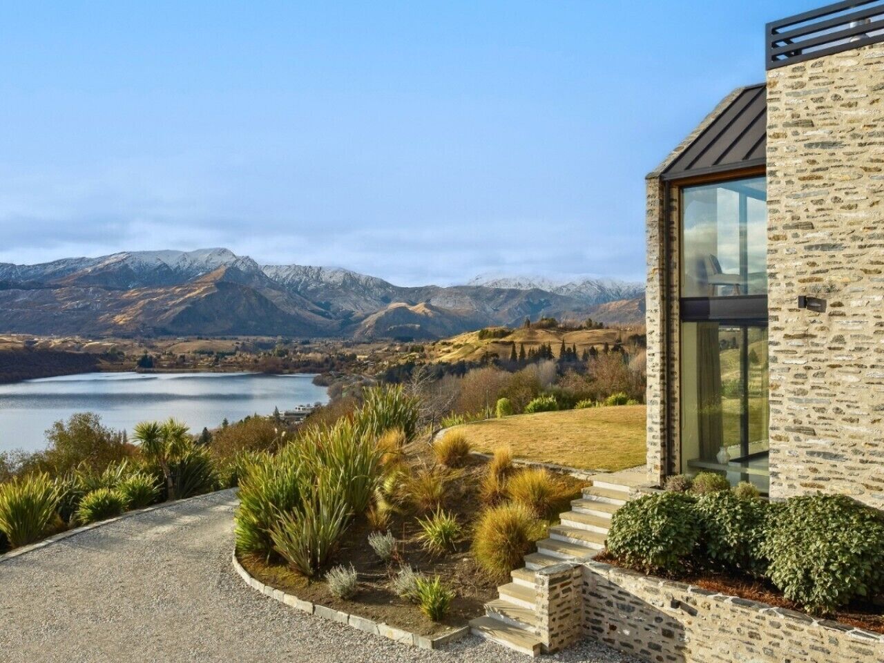 Una casa moderna con facciata in pietra, vialetto sterrato e vista su un lago e montagne.