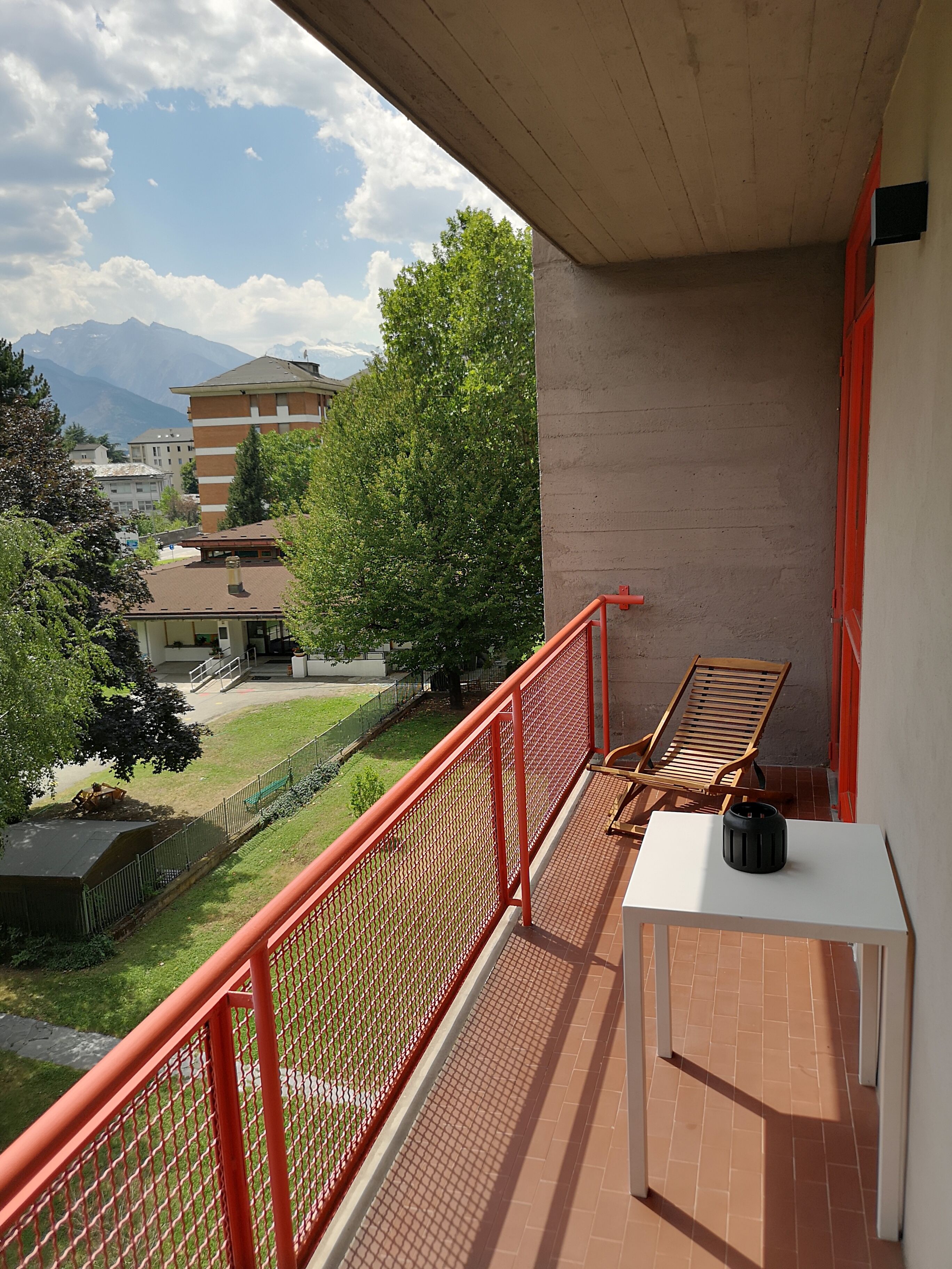 A balcony with a red railing, a wooden chair, and a small table with a black object on it.