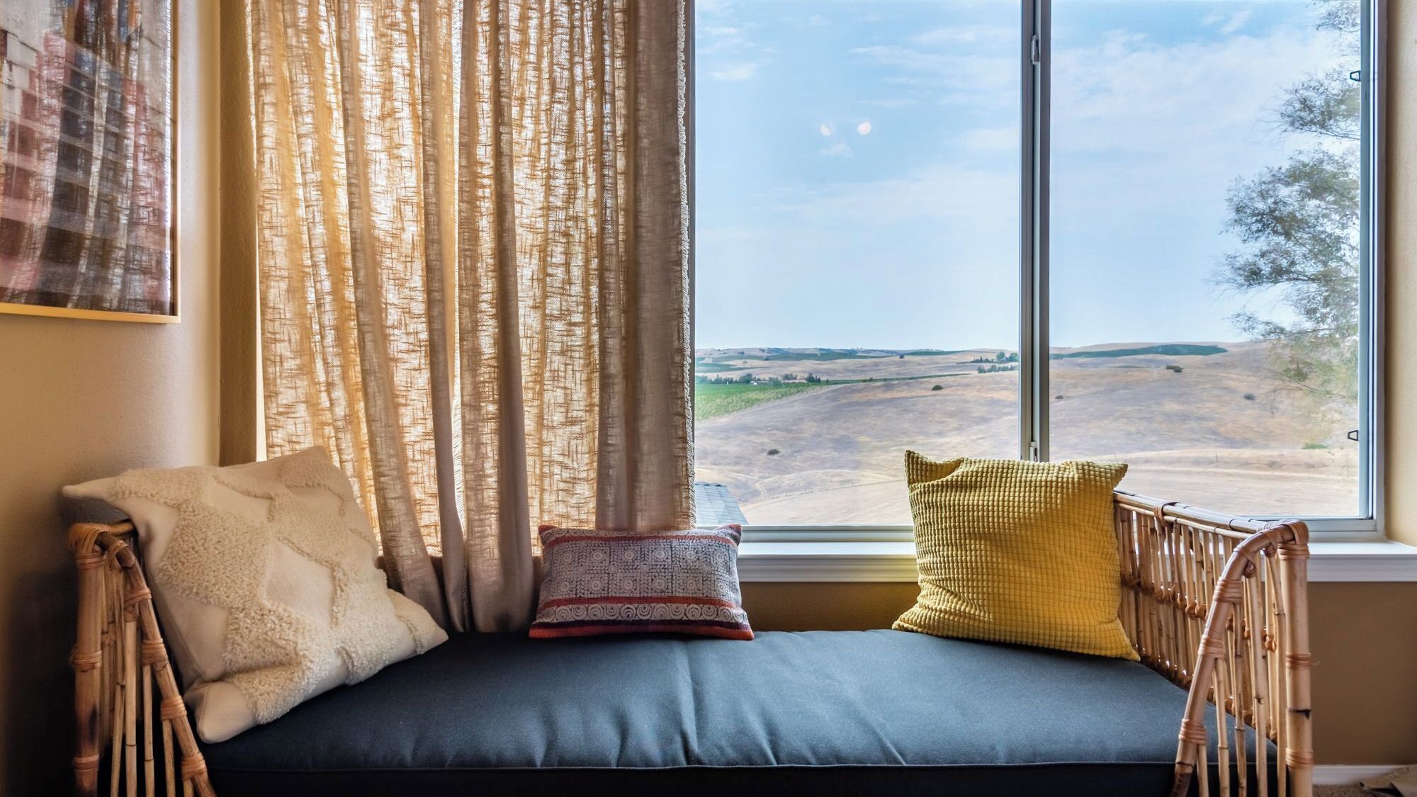 A room with a window overlooking a landscape, a cushioned bench with pillows, and a curtain.