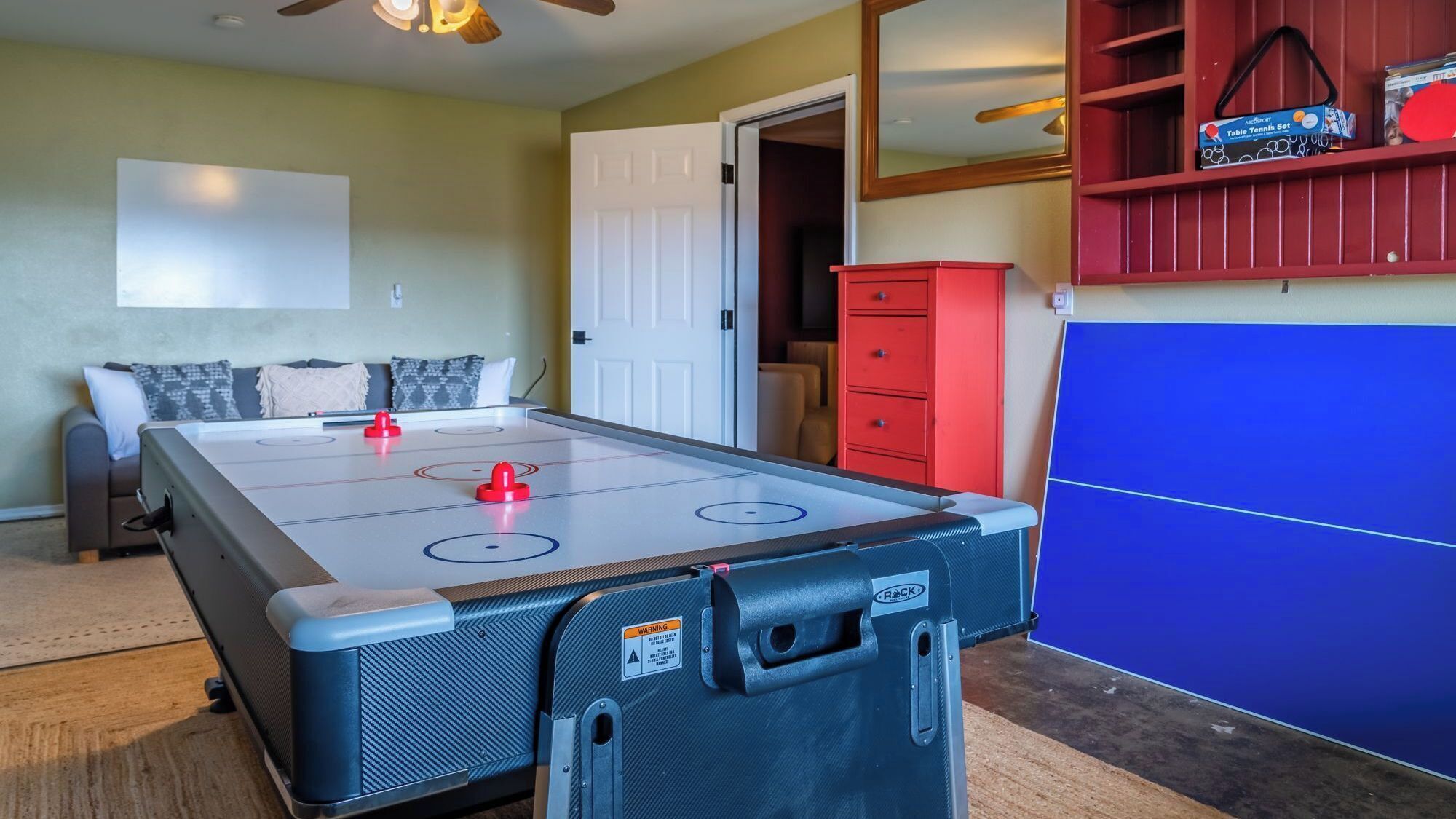 A room with a foosball table, a blue ping pong table, a red cabinet, and a sofa with cushions.