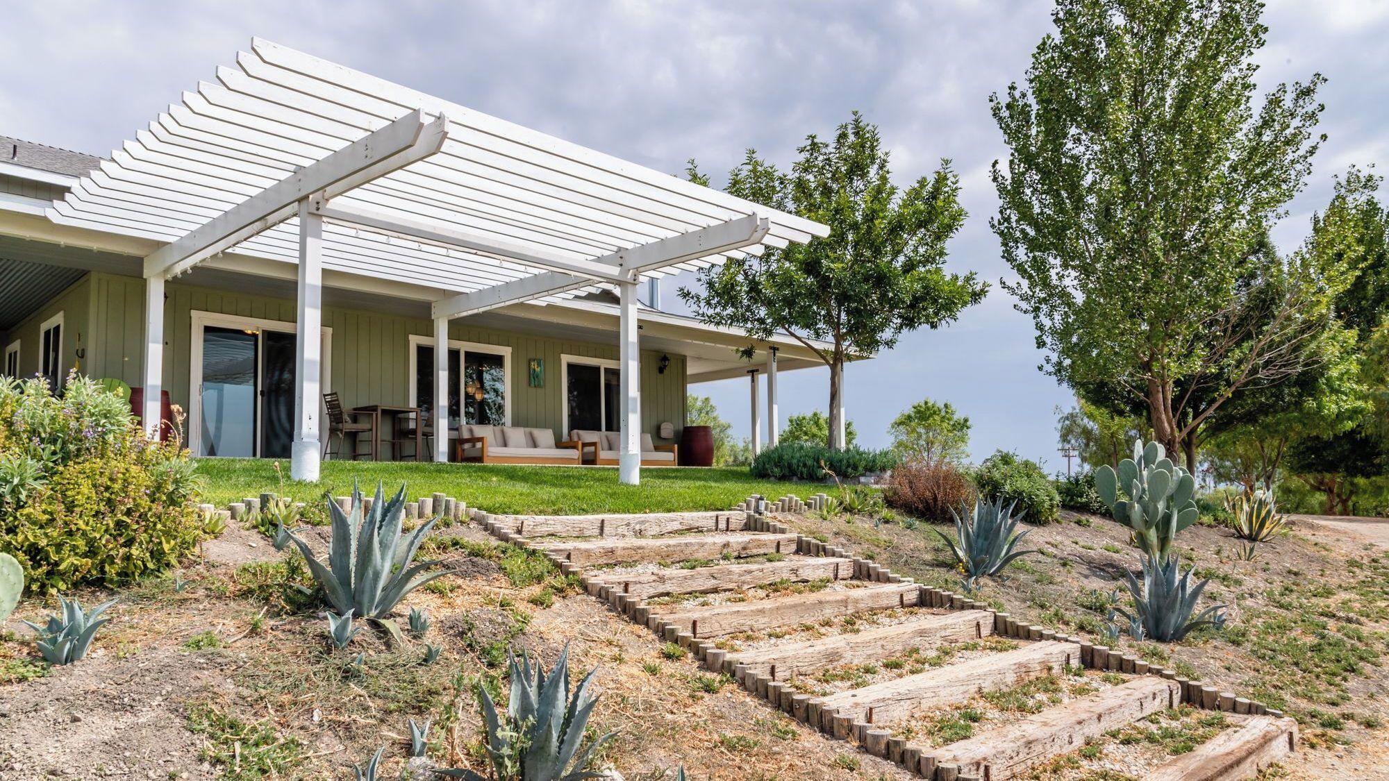 A house with a covered patio, a garden with various plants, and a wooden staircase leading up to the house.