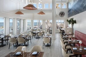 Un comedor amplio con varias mesas y sillas, ventanas grandes que ofrecen vista al mar, y lámparas colgantes.