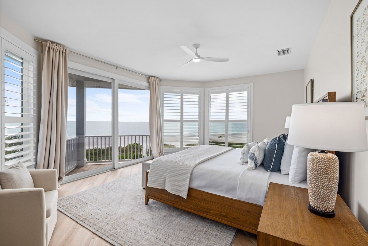Ein Schlafzimmer mit einem Bett, einem Nachttisch, einer Lampe, einem Deckenventilator und Blick ins Freie durch große Fenster.