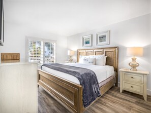 Ein Schlafzimmer mit einem hölzernen Bett, Nachttischen, einem Fenster mit Jalousien und gerahmten Bildern an der Wand.