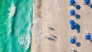 An aerial view of a beach with clear turquoise water, a sandy shore, and several rows of blue umbrellas and white lounge chairs. - South Seas Resort | 1BR Villa Near Captiva Landing (Captiva)