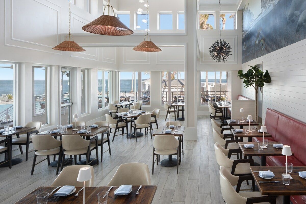 A spacious dining area with multiple tables and chairs, large windows offering a view of the sea, and hanging pendant lights.